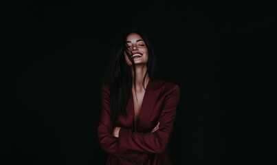 A joyful woman in a dark setting smiles brightly while wearing a stylish deep red blazer and crossing her arms