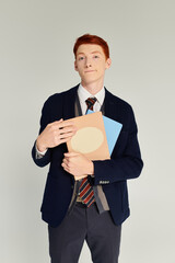 A young man with red hair stands elegantly in a blazer, showcasing a fashionable winter style while holding folders.