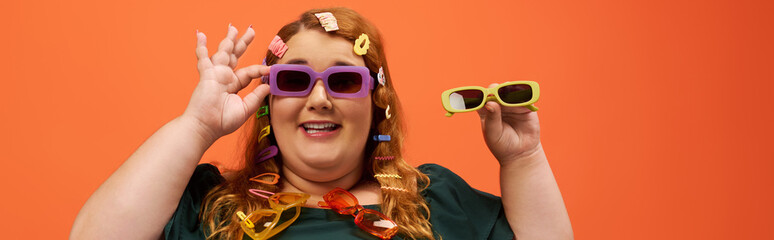 A cheerful plus size woman with stylish clips in her hair displays two pairs of trendy sunglasses.