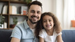 © halo - Happy father and daughter sitting on the couch in their living room, smiling at the camera.