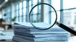 © Valeryi - a stack of papers lying on a desk in an office setting. In the foreground is a magnifying glass that magnifies part of the stack of papers