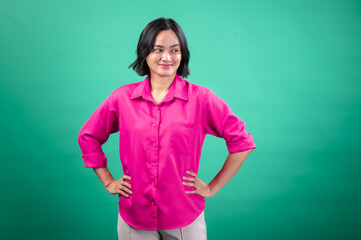 An Asian woman in a bright pink button-up shirt stands confidently with her hands on her hips against a solid green background, projecting a friendly and assertive expression