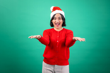 An Asian woman wearing a red sweater and Santa hat, smiling widely with one hand raised in a welcoming gesture, standing against a green background. She exudes festive joy and holiday cheer