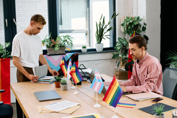 Two men sort paperwork in a lively, plant filled space adorned with pride flags.