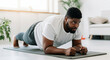 © Anna - Focused plus-size Black man practicing plank exercise on yoga mat in bright living room
