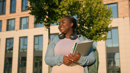 African American woman university college campus student girl schoolgirl with books schoolbag headphones walking in city walk waving hello hand greeting hi showing two fingers peace gesture outdoors