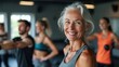 © SerPak - A mature woman smiles confidently while engaging in a fitness class alongside younger participants. The setting features a contemporary gym atmosphere with large windows and an encouraging energy amon