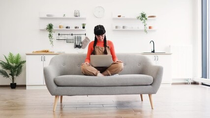 Wall Mural - Caucasian woman sitting in lotus pose meditating during remote work at home with laptop on sofa. Bright and modern living space with calming atmosphere.