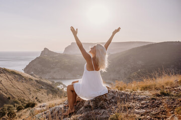 Wall Mural - Happy woman is sitting on a hillside, wearing a white dress. She is surrounded by a beautiful landscape, with a body of sea in the background. Concept of peace and happiness.