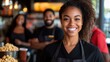 © Pinklife - A cheerful woman in a black apron stands at a popcorn counter, smiling with two colleagues in background, creating a friendly and welcoming atmosphere.
