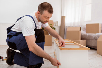 Fototapete - Professional Middle-Aged Carpenter Man Repairing Shelf Using Screwdriver, Assembling Furniture Indoors, Wearing Blue Coverall. Carpentry Service, Furniture Installation Concept