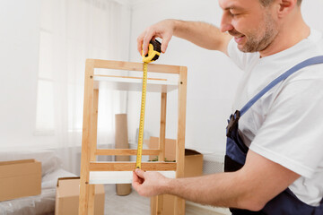 Fototapete - Carpentry, Furniture Assembling. Male Carpenter In Coverall Making Furniture Measuring Cabinet Shelf With Tape Standing Indoors. Craftsman Service Concept. Cropped, Selective Focus