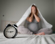 © Михаил Решетников - Caucasian pregnant woman sits on the bed and suffers from insomnia. Alarm clock in the foreground.