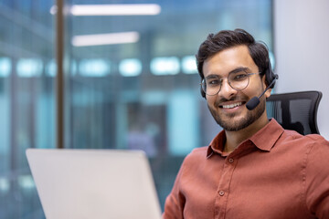 Wall Mural - Hispanic businessman wearing headset working on laptop in office setting. Confident smile, brown shirt, glasses, engaging with professionalism. Bright, modern background. Business and communication.