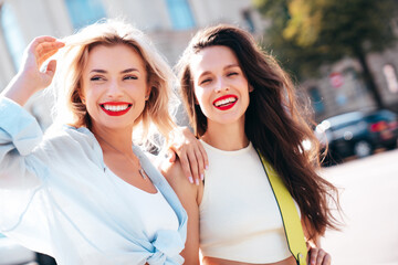 Wall Mural - Two young beautiful smiling female in trendy summer clothes. Sexy carefree women posing in the street. Positive models having fun, hugging in sunny day. With bright makeup and red lips