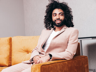 Handsome smiling hipster model. Unshaven Arabian man dressed in elegant beige suit . Fashion male with long curly hairstyle posing at home. sitting on yellow sofa