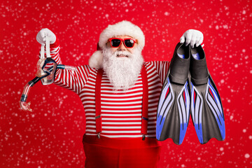 Photo of grandfather grey beard hold blue diving gear mask flippers smile wear santa claus x-mas costume suspenders sunglass striped shirt cap isolated red color background