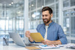 © Liubomir - Man happily opening envelope at desk with laptop in modern office, illustrating positive emotion, communication, and work success. Bright setting with stylish decor.
