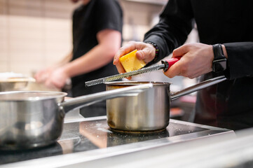 Wall Mural - A close-up of hands grating cheese over a pot on a stove, with another person cooking in the background. The kitchen scene is warm and inviting, showcasing culinary preparation and teamwork