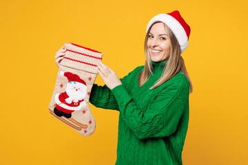 Wall Mural - Side view merry young woman wear green casual sweater Santa hat posing hold decorative stocking look camera isolated on plain yellow background. Happy New Year, celebration Christmas holiday concept.