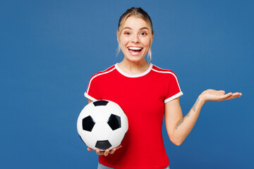 Young shocked surprised fun overjoyed woman fan wear basic red t-shirt cheer up support football sport team hold in hand soccer ball watch tv live stream look camera isolated on plain blue background.