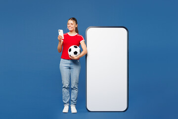 Full body young woman wear red t-shirt cheer up support football sport team hold soccer ball watch tv live stream big blank screen mobile cell phone use smartphone isolated on plain blue background.