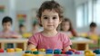 © KN Studio - Toddler's Curious Gaze: A captivating portrait of a toddler girl engrossed in colorful building blocks, surrounded by a blurred backdrop of other children in a playful classroom setting.