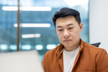 Wall Mural - Asian man businessman at desk in modern office setting wearing orange jacket focused on work. Large glass windows create bright atmosphere. Represents professionalism and dedication.