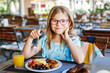 © Irina Schmidt - Adorable little girl having breakfast at resort restaurant. Happy preschool or school child eating healthy food, vegetables and eggs in the morning.