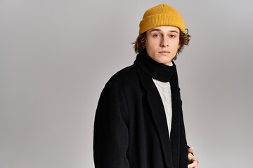 A young man with a charming expression stands in a studio, wearing a sleek black coat and a bright yellow beanie.