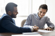 © fizkes - Happy young multiethnic male partners signing agreement, contract after negotiation, business discussion at work table. Seller pointing at paper for buyer, client setting signature