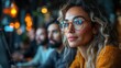 © Kalivmasa - A young woman with curly hair and glasses sits at a table with other people in the background. She looks intently at something in the distance, and her expression is thoughtful and focused.