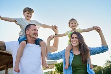 Wall Mural - Parents, children and airplane on shoulders, playful and happy with holding hands, love and summer. Mother, father and kids with play, flight and piggy back with smile in garden with game in Spain