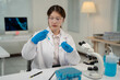 © Tj - Young scientist wearing lab coat and gloves comparing two test tubes with blue liquid while sitting at desk in modern laboratory with microscope and computer