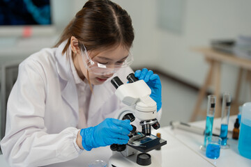 Female scientist wearing lab coat and gloves carefully adjusting microscope, examining sample on slide in modern laboratory, surrounded by test tubes and beakers containing blue liquid