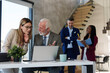 © Dorde - Strategic discussion unfolds as a focused Caucasian businessman works on his laptop, with colleagues engaging in the background. Professional attire and diverse teamwork evident in a modern office.