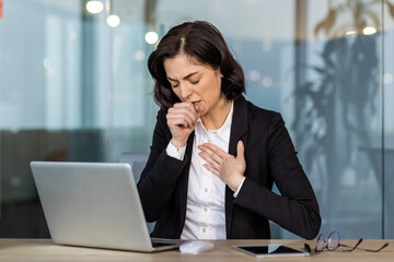 Poster - Young businesswoman wearing suit coughing while working on laptop in office setting. Health issues or discomfort during worktime illustrating common workplace challenges.