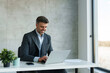 © Dorde - A mid adult Caucasian man dressed in business attire focuses on his laptop, engaged in work at a bright office with a minimalist style.