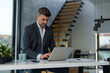 © Dorde - A focused, mid adult Caucasian businessman in a tailored suit engages with his laptop at a sleek desk in a well-lit contemporary office setting.
