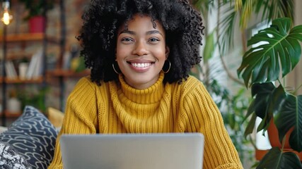 Aufkleber - A cheerful young woman wearing a bright yellow sweater smiles at the camera as she works on her laptop in a warm, plant-filled living room during the afternoon.