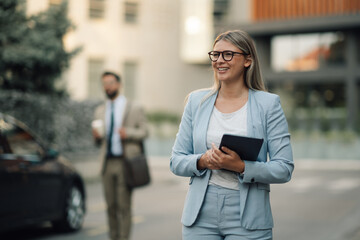 Smiling businesswoman holding tablet walking in city with businessman in background