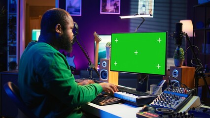 Poster - Musician operates on midi controller piano keys next to mockup screen, producing new songs with musical instruments and audio gear in home studio. Artist works with green screen. Camera B.