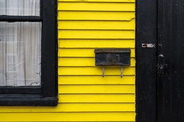 A bright and vibrant yellow exterior wall to a vintage wooden building. There's a black wood storm door with a hinge, a black mailbox, and a closed glass window in the horizontal wood siding frame. 