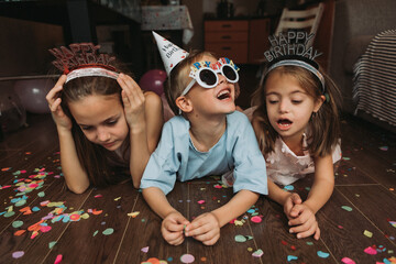 Three small children celebrate their birthday at home, lying on the wooden floor with confetti