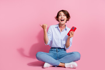 Wall Mural - Full body portrait of charming impressed lady sit floor hold smart phone look indicate finger empty space isolated on pink color background