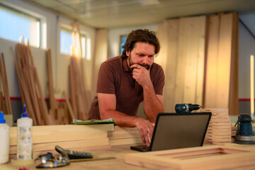 Owner of small business and a woodworking shop, using a laptop while reviewing documents.Dressed in work suit, stands at a workbench with wooden planks and tools around him.