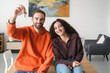 © Studio Marmellata - A cheerful young couple sitting together at a table, with the man in an orange hoodie holding up a set of keys, symbolizing a fresh beginning or new home in a brightly lit, modern room.