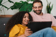 © Studio Marmellata - A relaxed couple sitting closely on a couch, engaged with a tablet, in a cozy living room adorned with indoor plants, creating a warm and welcoming atmosphere in a shared moment.