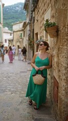 Wall Mural - Young hispanic woman in a green dress leaning against a stone wall in a picturesque street of valldemossa, mallorca, spain outdoors with mountains in the background.