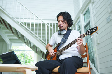 Young Asian Japanese man with headphones practicing bass guitar indoors, focusing on digital tablet screen for music lessons in a modern home interior with staircase in background.